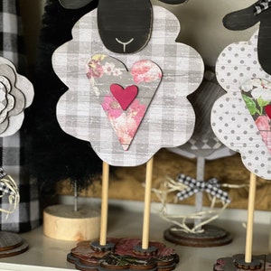 May include: Three black and white sheep figurines with floral hearts. The sheep are standing on wooden bases with floral designs. The sheep are a farmhouse style decor.