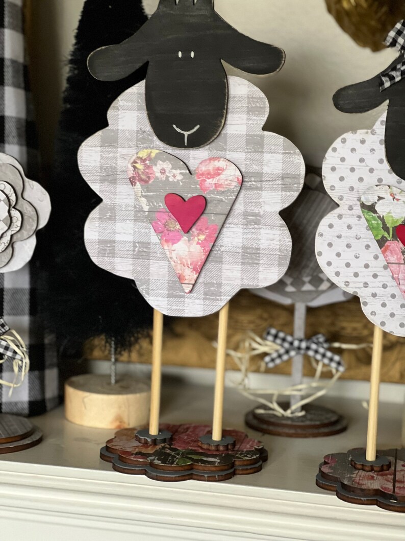 May include: Three black and white sheep figurines with floral hearts. The sheep are standing on wooden bases with floral designs. The sheep are a farmhouse style decor.