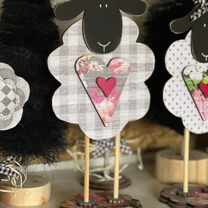 May include: Black and white sheep figurine with a pink heart and floral pattern. The sheep is standing on a wooden base with a floral design.