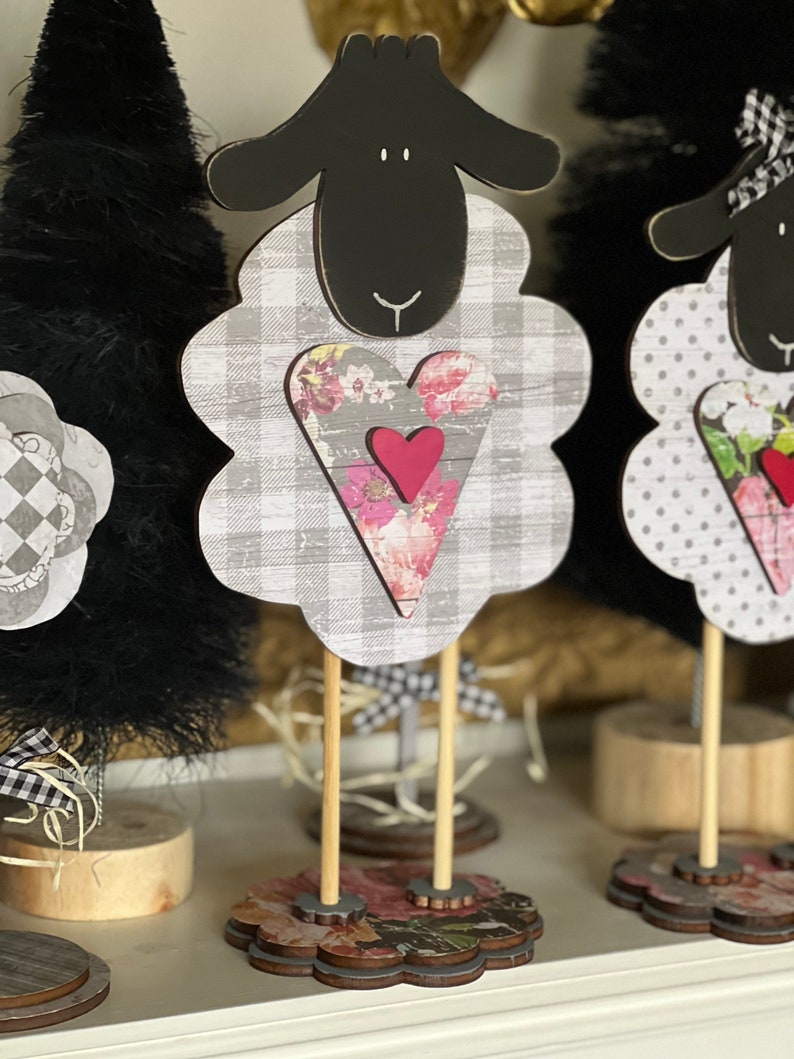 May include: Black and white sheep figurine with a pink heart and floral pattern. The sheep is standing on a wooden base with a floral design.