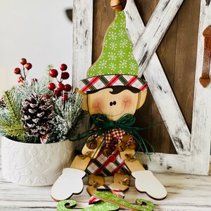 May include: A wooden elf figurine with a green hat, red and green plaid shirt, and white mittens. The elf is sitting on a wooden surface with a green and white patterned sled.