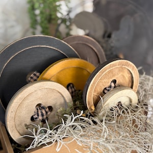 May include: A wooden crate filled with decorative wooden spools in various colors, including black, brown, and yellow. Each spool features a plaid fabric accent in the center. The crate is filled with dried plants.