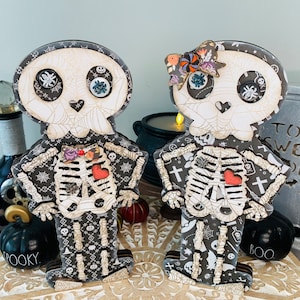 May include: Two wooden skeleton figures with black and white patterned clothing. The skeleton on the left is wearing a red heart and the skeleton on the right is wearing a pink heart. Both skeletons have black eyes and a spider web design on their heads.