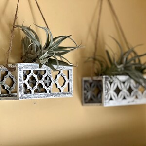 May include: Three white wooden hanging planters with intricate cut-out designs. Each planter is filled with a green air plant and hangs from a string.