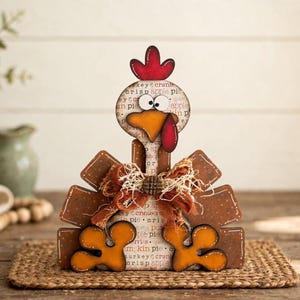 Pu&ograve; includere: Una statuetta decorativa a forma di tacchino in legno, con cresta rossa, becco arancione e volto bianco con motivi testuali neri. Il corpo presenta piume di coda marroni smerlate, zampe arancioni e un fiocco in tessuto arancione sfilacciato.