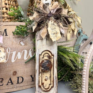 May include: A rustic wooden door knob hanger with a distressed white paint finish. The hanger features a brown fabric bow with a brown button in the center. A small tag with the text "He is the God of all comfort" is attached to the bow. The hanger is perfect for adding a touch of farmhouse charm to your home decor.