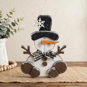 Op de afbeelding: Een houten sneeuwpopfiguur met een zwarte hoge hoed met een witte sneeuwvlok, een feloranje wortelneus en een zwart-wit geblokte strik. De sneeuwpop heeft twee bruine knopen op het lichaam en bruine takken als armen.