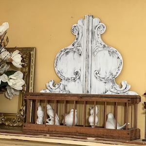 Può includere: Una gabbia per uccelli in legno rustico con un pezzo decorativo in legno dipinto di bianco invecchiato in cima. La gabbia per uccelli contiene cinque uccelli in porcellana bianca all'interno. La gabbia per uccelli è appoggiata su uno scaffale con un quadro incorniciato in oro e un arrangiamento di fiori bianchi.