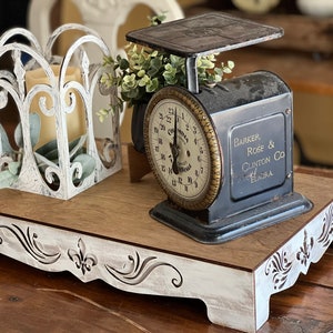 May include: A white wooden tray with decorative cutouts features a vintage black scale with a gold face and a white metal candle holder. The scale reads "Barker, Rose & Clinton Co. Elmira".