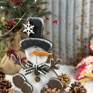 Op de afbeelding: Een platte houten sneeuwpopdecoratie met een zwarte hoge hoed met een wit sneeuwvlokaccent, een oranje wortelneus, een zwart-witte buffalo ruitjesstrik, twee bruine knopen en donkerbruine takvormige armen en voeten.