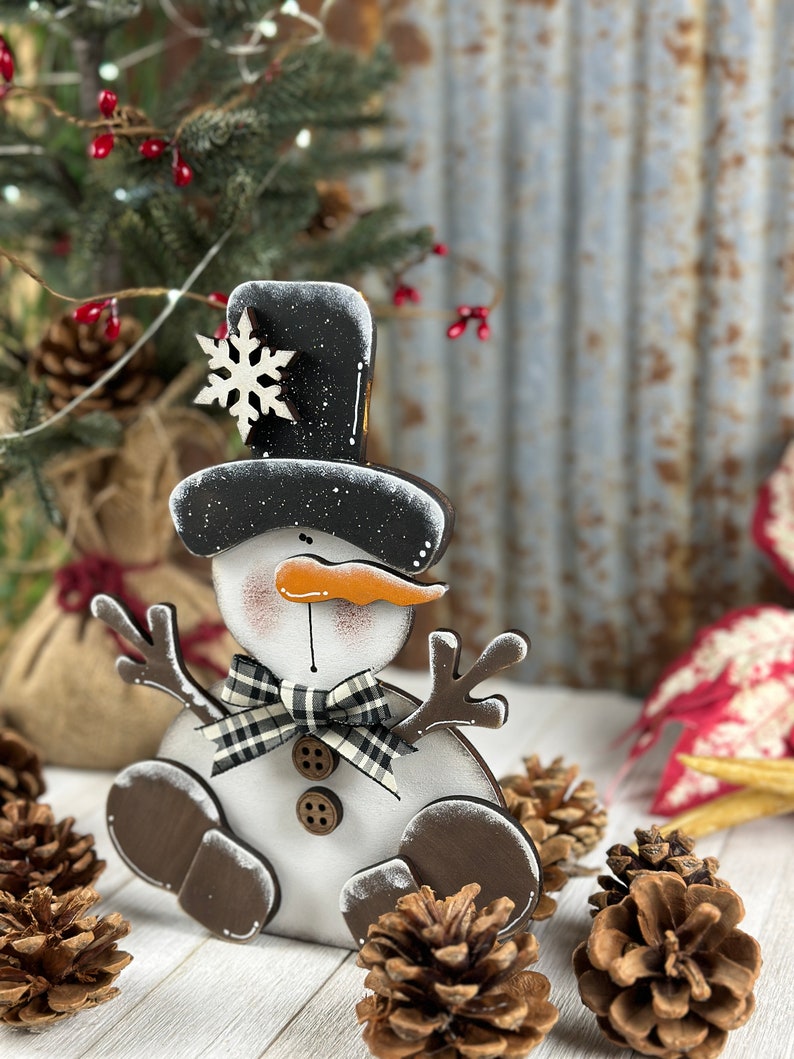 Op de afbeelding: Een platte houten sneeuwpopdecoratie met een zwarte hoge hoed met een wit sneeuwvlokaccent, een oranje wortelneus, een zwart-witte buffalo ruitjesstrik, twee bruine knopen en donkerbruine takvormige armen en voeten.