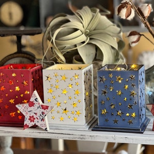 Puede incluir: Tres portavelas de madera con recortes en forma de estrella. Los portavelas son rojo, blanco y azul. Cada portavelas tiene una cadena de luces en su interior.