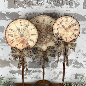 May include: Three vintage-style clock faces on rusty metal stands with brown and white checkered ribbon bows. The clocks have Roman numeral markings and a distressed, aged look. The text "Pickitz Crafts" is visible at the top of the image.