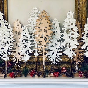 Puede incluir: Un conjunto de siete árboles de madera recortados en blanco y marrón con un tema invernal. Los árboles están dispuestos en una fila sobre una superficie de madera con bayas rojas y piñas esparcidas a su alrededor.
