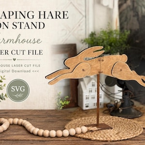 May include: A wooden leaping hare figure mounted on a thin metal stand. The text LEAPING HARE ON STAND Farmhouse LASER CUT FILE and SVG is visible. The hare is composed of three connected wooden pieces with visible joints and a light brown finish.