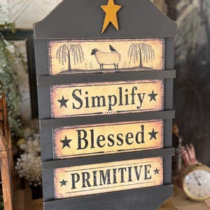 May include: A black wooden wall hanging with a gold star on top. The sign has three panels with the words "Simplify", "Blessed", and "Primitive" in a vintage font. The top panel features a sheep and bird illustration.