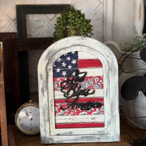 Könnte beinhalten: Ein weißer Holzrahmen mit gewölbtem Oberteil. Im Inneren befindet sich ein patriotisches Design mit der amerikanischen Flagge und floralem Stoff. Die Worte "God Bless America" sind in schwarzer Schrift ausgeschnitten. Ein dekoratives Stück.