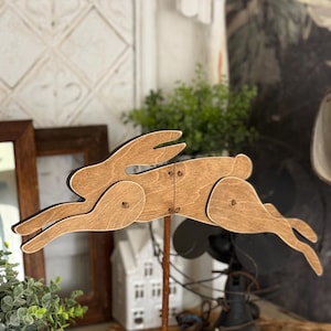 May include: A wooden rabbit figurine in a jumping pose, painted in a light brown color, mounted on a rusty metal rod. The rabbit is made of multiple pieces of wood, with visible joints and holes for attaching the pieces.