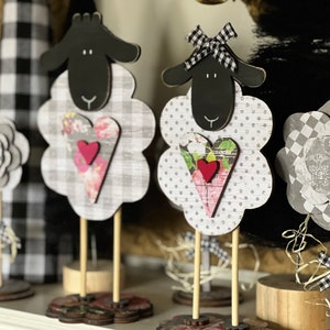 May include: Three black and white sheep figurines with hearts and bows. The sheep are made of wood and have a rustic farmhouse style.