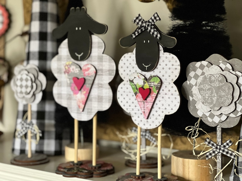 May include: Three black and white sheep figurines with hearts and bows. The sheep are made of wood and have a rustic farmhouse style.