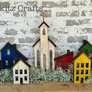 Pode incluir: Um conjunto de casas de madeira pintadas em várias cores, incluindo azul, branco, verde, vermelho e amarelo. As casas estão dispostas em uma fileira sobre uma superfície de madeira com vegetação na frente delas. A maior casa é uma igreja com uma cruz no topo. O texto "Pickitz Crafts" é visível na parte superior da imagem.