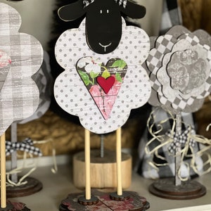 May include: Three wooden sheep figurines with black and white checkered fabric and a heart-shaped design. The sheep on the right has a black head and a white body with black polka dots. The sheep in the middle has a black head and a white body with gray polka dots. The sheep on the left has a gray and white checkered body. All three sheep have wooden legs and stand on a wooden base.