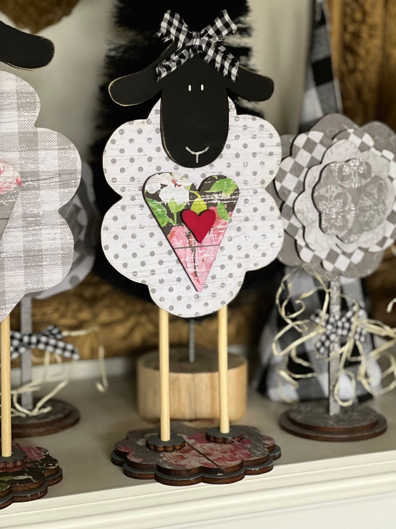 May include: Three wooden sheep figurines with black and white checkered fabric and a heart-shaped design. The sheep on the right has a black head and a white body with black polka dots. The sheep in the middle has a black head and a white body with gray polka dots. The sheep on the left has a gray and white checkered body. All three sheep have wooden legs and stand on a wooden base.