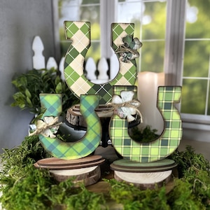 May include: Three wooden horseshoes decorated with green and yellow plaid and argyle patterns. Each horseshoe has a small green shamrock attached to it. The horseshoes are sitting on wooden rounds.