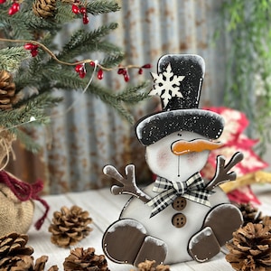 Op de afbeelding: Een houten sneeuwpopdecoratie met een zwarte hoge hoed met een wit sneeuwvlokaccent, een wortelvormige neus, twee bruine knopen, een zwart-wit geblokte strik en bruine, takachtige armen en afgeronde voeten.