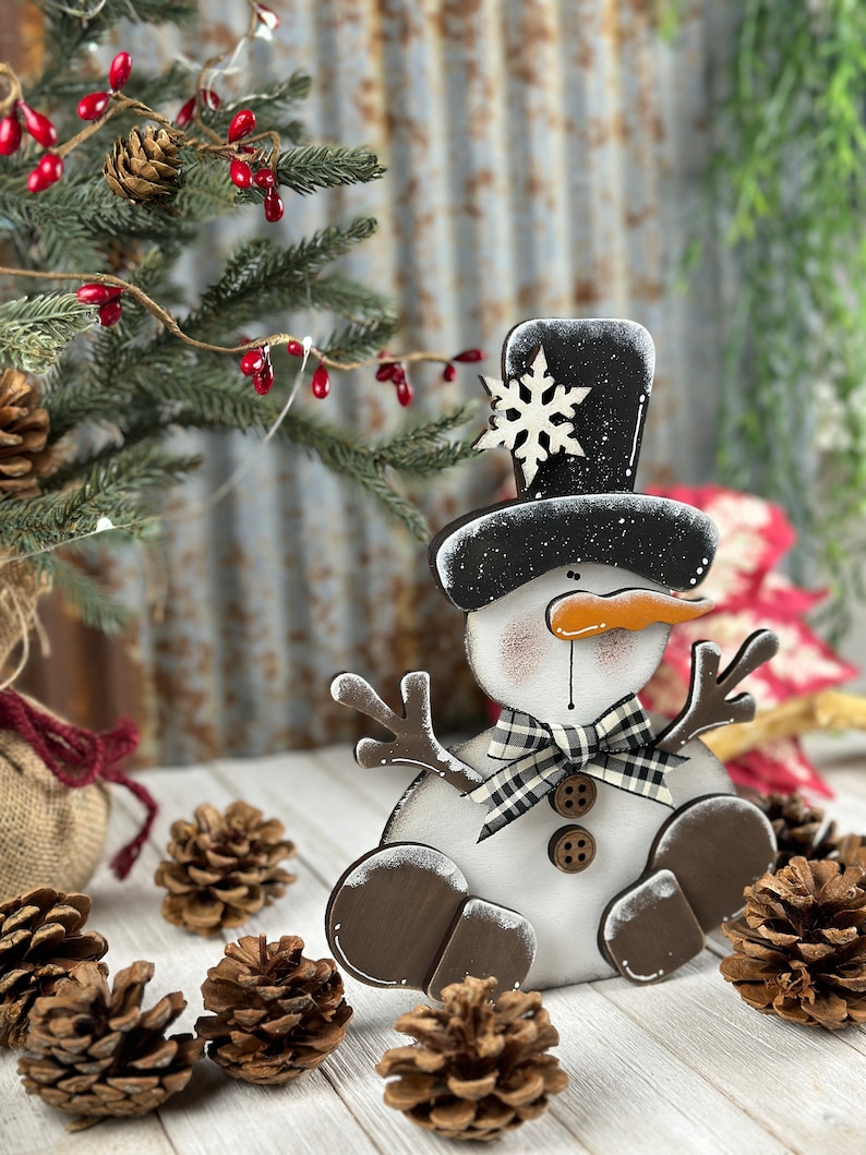 Op de afbeelding: Een houten sneeuwpopdecoratie met een zwarte hoge hoed met een wit sneeuwvlokaccent, een wortelvormige neus, twee bruine knopen, een zwart-wit geblokte strik en bruine, takachtige armen en afgeronde voeten.