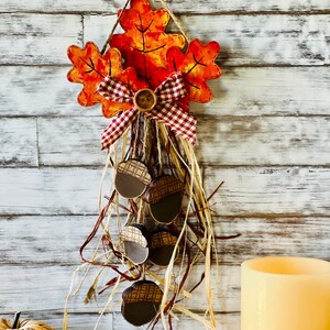May include: A fall-themed door hanger with a red and white checkered bow and a brown button. The hanger features painted orange, red, and yellow leaves and a cluster of brown acorns hanging from the bottom.