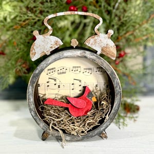May include: A vintage-style alarm clock with a distressed finish, featuring a red cardinal bird perched in a nest made of brown straw. The clock face is a sheet of music with musical notes printed on it.