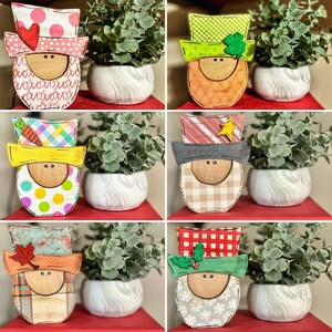 May include: Six wooden gnome ornaments with different hats and facial expressions. Each gnome is painted with various patterns and colors, including red, green, blue, and yellow. The gnomes are displayed on a red surface with small potted plants.