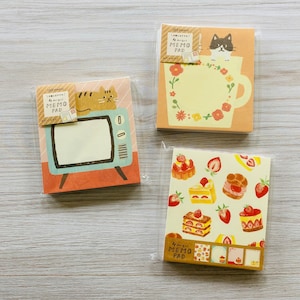 May include: Three memo pads in plastic packaging. One pad features a cat on a retro TV, another has a cat in a teacup with flowers, and the third displays strawberry pastries. Each pad has 100 sheets and the words "MEMO PAD".