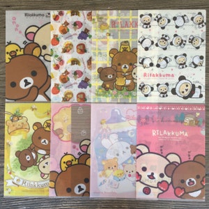 May include: A selection of Rilakkuma cellophane bags with diverse designs. The bags display illustrations of bears, chicks, and other characters in various poses and settings. Some bags have text, and the colour palette includes yellow, pink, and white.