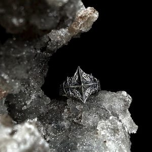 May include: A silver ring featuring a prominent, sharp-pointed star or compass-like design at the center. The band is decorated with intricate, raised knotwork patterns that extend across the surface, creating a textured and detailed appearance.