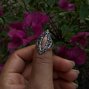 May include: A silver-colored ring with an ornate, gothic-inspired design. The ring features an elongated oval center and intricate detailing, held up by a hand against a backdrop of vibrant purple flowers and green foliage. The ring's design is symmetrical and detailed.