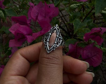 May include: A silver-colored ring with an ornate, gothic-inspired design. The ring features an elongated oval center and intricate detailing, held up by a hand against a backdrop of vibrant purple flowers and green foliage. The ring's design is symmetrical and detailed.