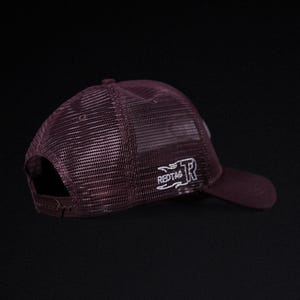 May include: Maroon trucker hat with a mesh back and a curved brim. The hat features the embroidered text "REDTAG" and a stylized "R" logo in white on the side. Adjustable snap closure.