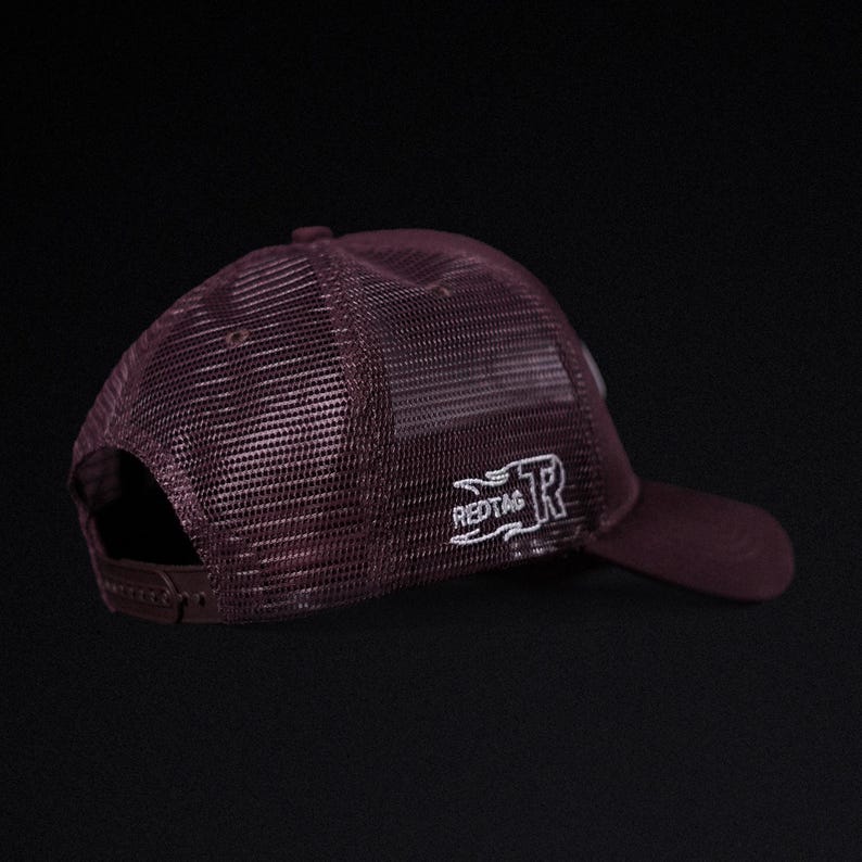 May include: Maroon trucker hat with a mesh back and a curved brim. The hat features the embroidered text "REDTAG" and a stylized "R" logo in white on the side. Adjustable snap closure.
