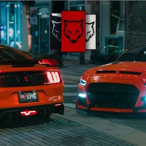 May include: Two orange Ford Mustang cars parked on a city street at night. The car on the left has its taillights on and the car on the right has its headlights on. The car on the left has a Florida license plate that reads "BOPHG".