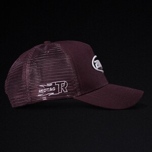 May include: A burgundy trucker hat with a mesh back and a curved brim. The hat features white embroidered logos on the front and side, including the text "REDTAG". The hat is designed for casual wear.