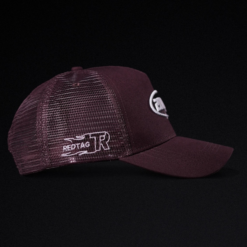 May include: A burgundy trucker hat with a mesh back and a curved brim. The hat features white embroidered logos on the front and side, including the text "REDTAG". The hat is designed for casual wear.