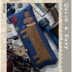 May include: A blue rectangular pincushion with a brown dachshund design, red stitching, and colorful pins. The text "Snag the Sausage Dog Pincushion" is at the bottom. The image also includes buttons, berries, and the text "Quick & Easy".