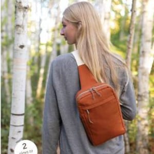 May include: A rust-colored sling bag with a white strap is worn over the shoulder. The bag has a zippered main compartment and a smaller front pocket. The text "SANDHILL" and "a compact hands-free sling style bag" are at the top. The text "2 views to sew!" is in a white circle.