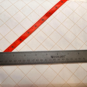 Puede incluir: Una superficie blanca con un patrón de cuadrícula de finas líneas doradas. Una cinta métrica roja se coloca en diagonal sobre la superficie. Una regla negra con marcas blancas se coloca horizontalmente en la parte inferior de la imagen. La regla está etiquetada como "WESTCOTT" y tiene marcas en pulgadas y centímetros.