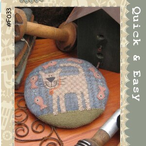 May include: A round pincushion with a light blue background features a beige cat design and pink fish accents. The pincushion is surrounded by a green border. The text "My Cat Claude Pincushion" is at the bottom.