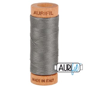 May include: A spool of gray Aurifil thread. The thread is wound around a cylindrical core with wooden ends. The top of the spool is stamped with "AURIFIL", and the bottom reads "MADE IN ITALY". The Aurifil logo is visible on the right.