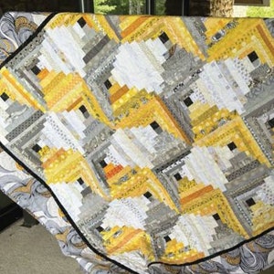 May include: A handmade quilt with a geometric pattern in shades of yellow, gray, and white. The quilt features a log cabin design with a black border and a patterned backing. The quilt is likely made of cotton or a similar fabric.