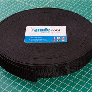 May include: A roll of black webbing material, likely for crafting or bag making. A business card with the text "byannie.com" rests on top. The background is a green cutting mat with grid lines.